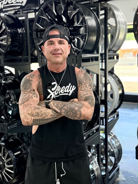 Man with extensive arm tattoos and black cap standing with arms crossed in front of display of custom black and silver wheels